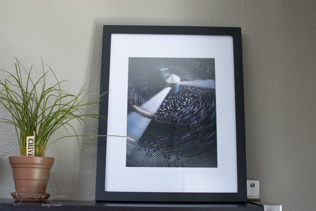 A framed photograph on a shelf next to a potted plant. The photo depicts a coffee roaster in action, with beans swirling inside. The frame is black and contrasts against the light gray wall.