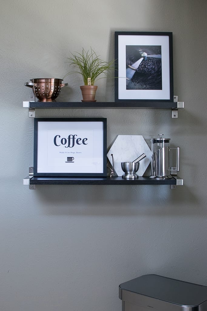 Free Printables Two black shelves on a gray wall hold a potted plant, framed photos, and coffee-related items, including a French press and a colander. One framed print reads "Coffee.