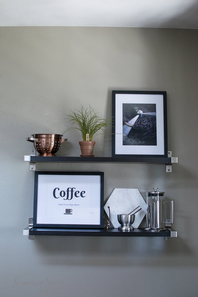 Two wall-mounted shelves on a light gray wall. Top shelf holds a copper colander, potted plant, and black-and-white photo. Bottom shelf displays a framed "Coffee" poster, a marble trivet, a coffee press, and a small metal cup and saucer.