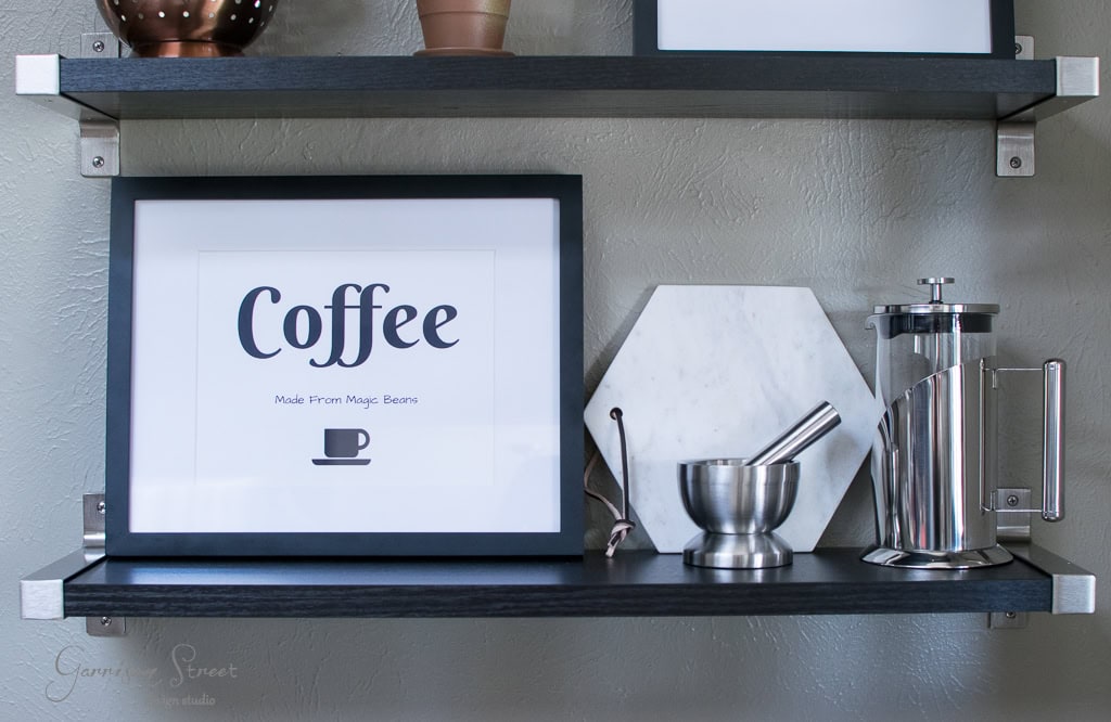 A kitchen shelf with a framed print saying "Coffee Made From Magic Beans," featuring a small coffee cup graphic. Next to it are a marble hexagon, a mortar and pestle, and a stainless steel French press.