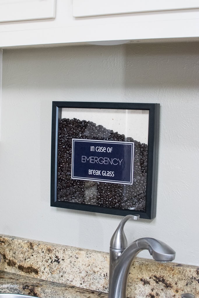 A framed shadow box filled with coffee beans hangs on a kitchen wall above a granite countertop. The frame has a sign that reads "In case of emergency, break glass." A silver kitchen faucet is visible below.