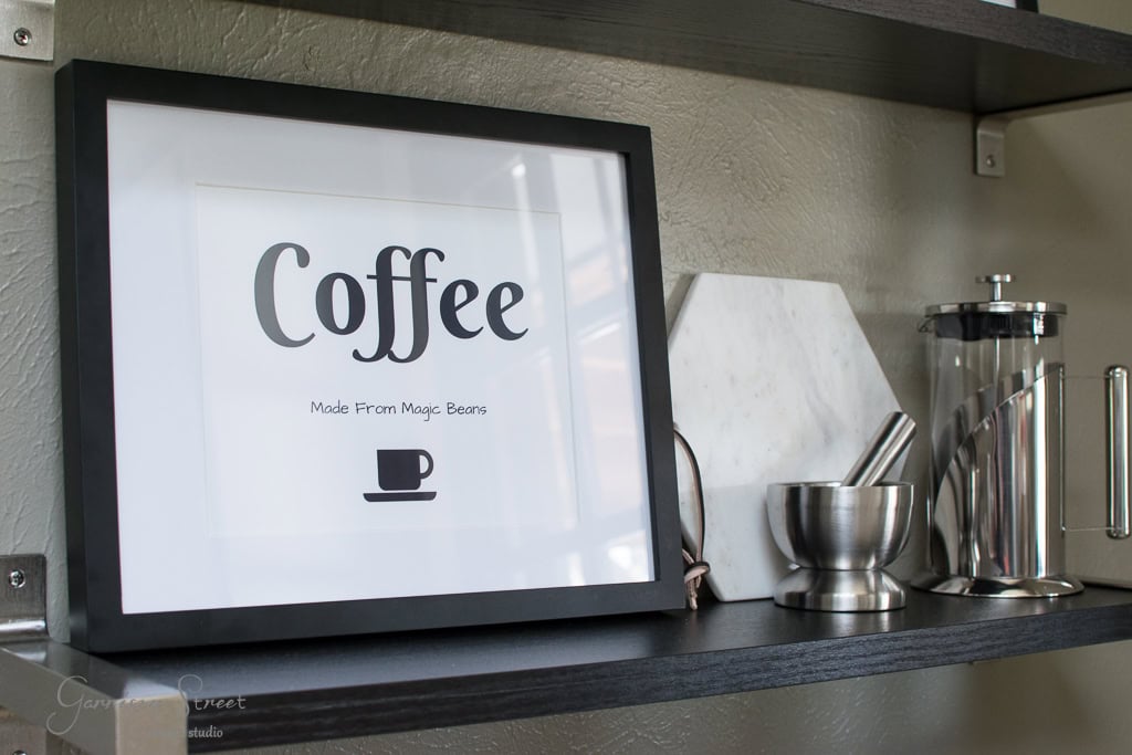 A framed sign on a shelf reads "Coffee Made From Magic Beans" with a coffee cup illustration beneath. Surrounding items include a metal mortar and pestle, a marble board, and a French press, all on a dark wooden shelf.