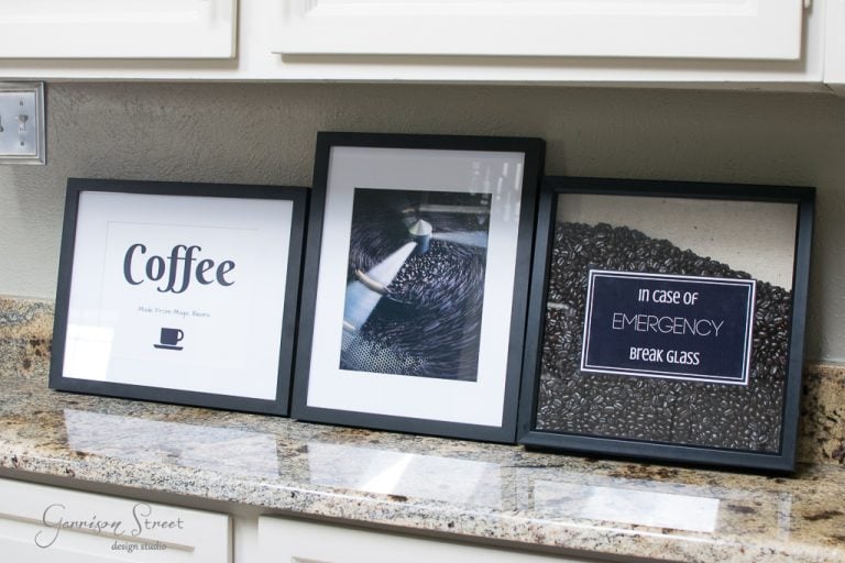 Three framed coffee-themed prints on a kitchen counter. The first says "Coffee Makes Magic Happen" with a cup icon, the middle features a close-up image of coffee beans, and the last reads "In Case of Emergency Break Glass" with beans behind glass.