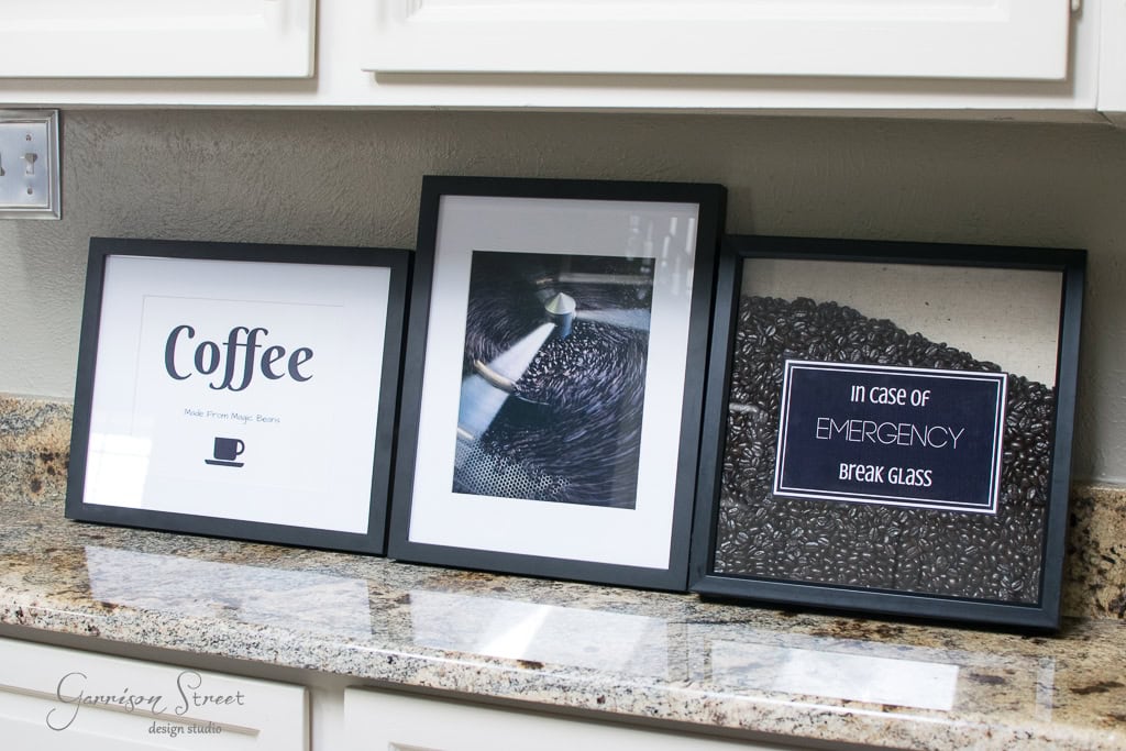Three framed coffee-themed prints on a kitchen counter. The first says "Coffee Makes Magic Happen" with a cup icon, the middle features a close-up image of coffee beans, and the last reads "In Case of Emergency Break Glass" with beans behind glass.