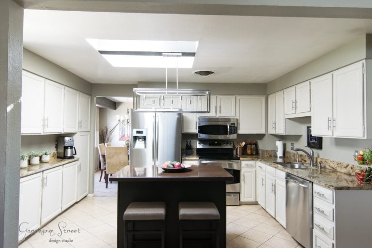 A modern kitchen with white cabinets, a stainless steel refrigerator, oven, and dishwasher. A central island with two stools features a fruit bowl. A skylight brightens the space, and a dining room is visible in the background.