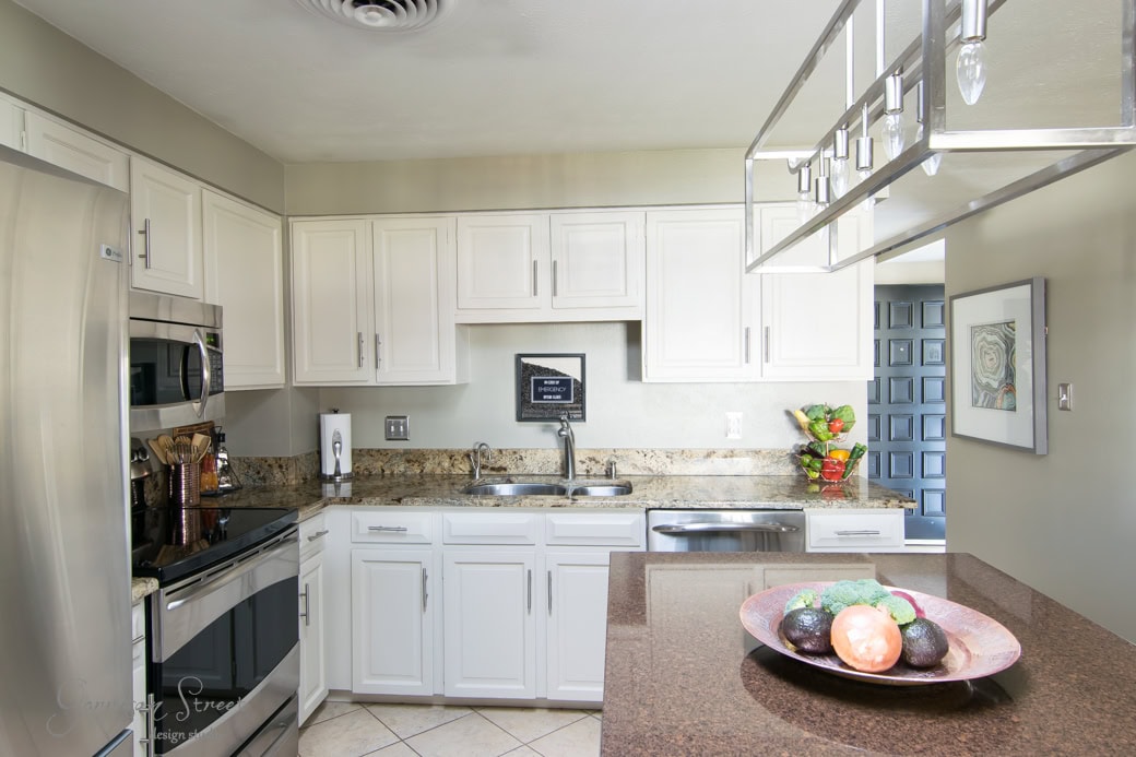 A modern kitchen with white cabinets, stainless steel appliances, and granite countertops. There's a fruit bowl on the island featuring avocados and an onion. Bright flowers are displayed near the window, adding a colorful touch.