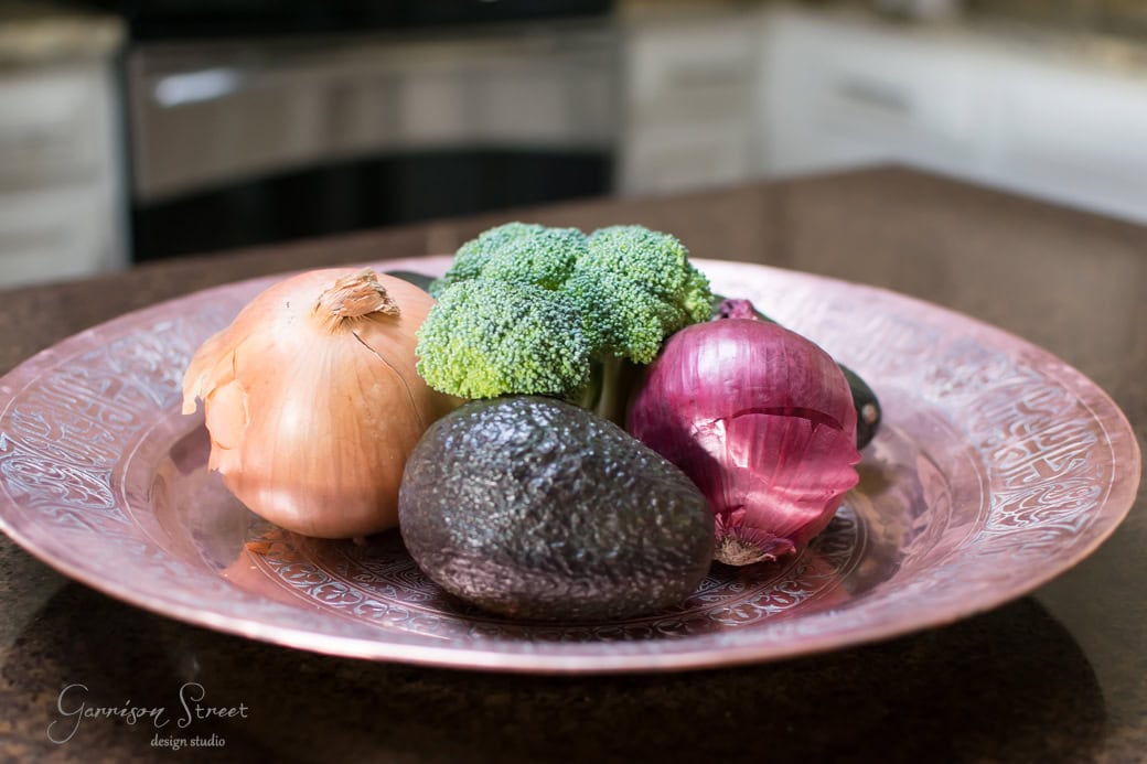 A copper plate on a kitchen counter holds fresh produce, including an onion, broccoli, an avocado, and a red onion.