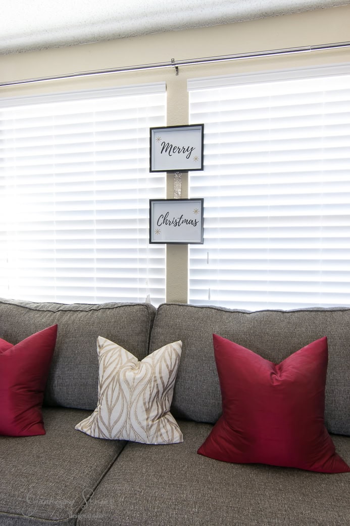 A gray sofa with two red pillows and one white pillow with a gold leaf pattern is in front of white blinds. Above the sofa, two framed signs hang vertically, reading "Merry" and "Christmas.