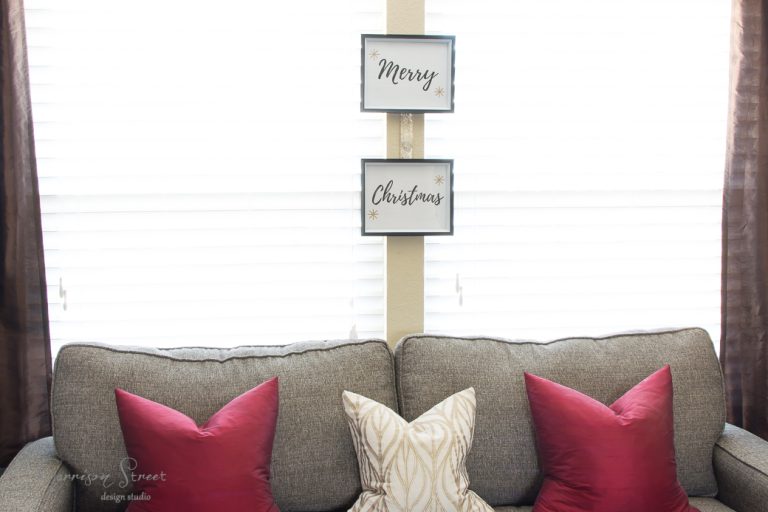 A cozy gray couch with two red pillows and a cream patterned pillow is in front of a window with white blinds. Above the couch, two framed signs hang vertically, reading "Merry" and "Christmas".