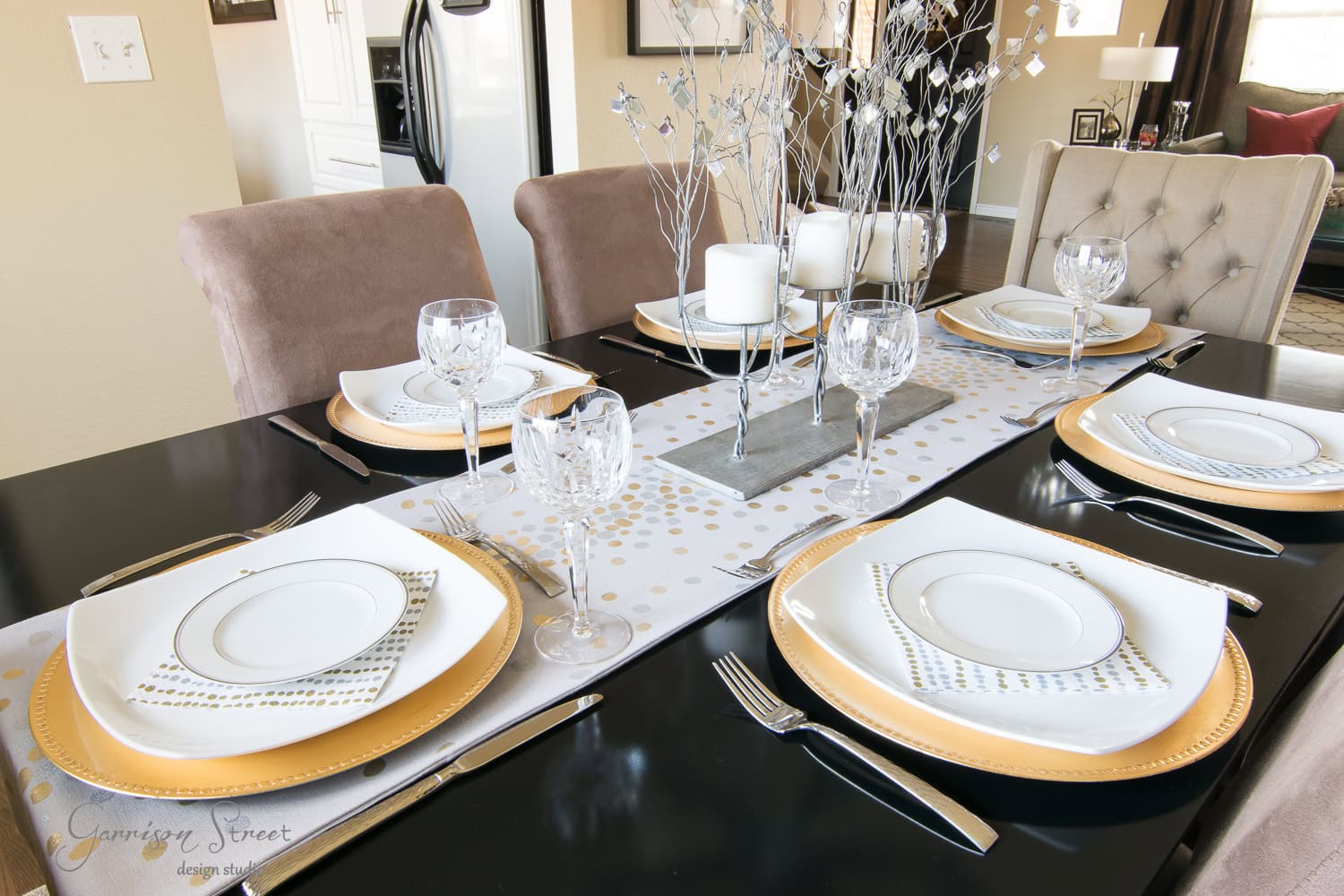 A dining table is elegantly set with gold and white dinner plates, crystal wine glasses, and silver cutlery. A festive table runner decorated with metallic dots runs down the center, topped with white candles and a silver branch centerpiece.