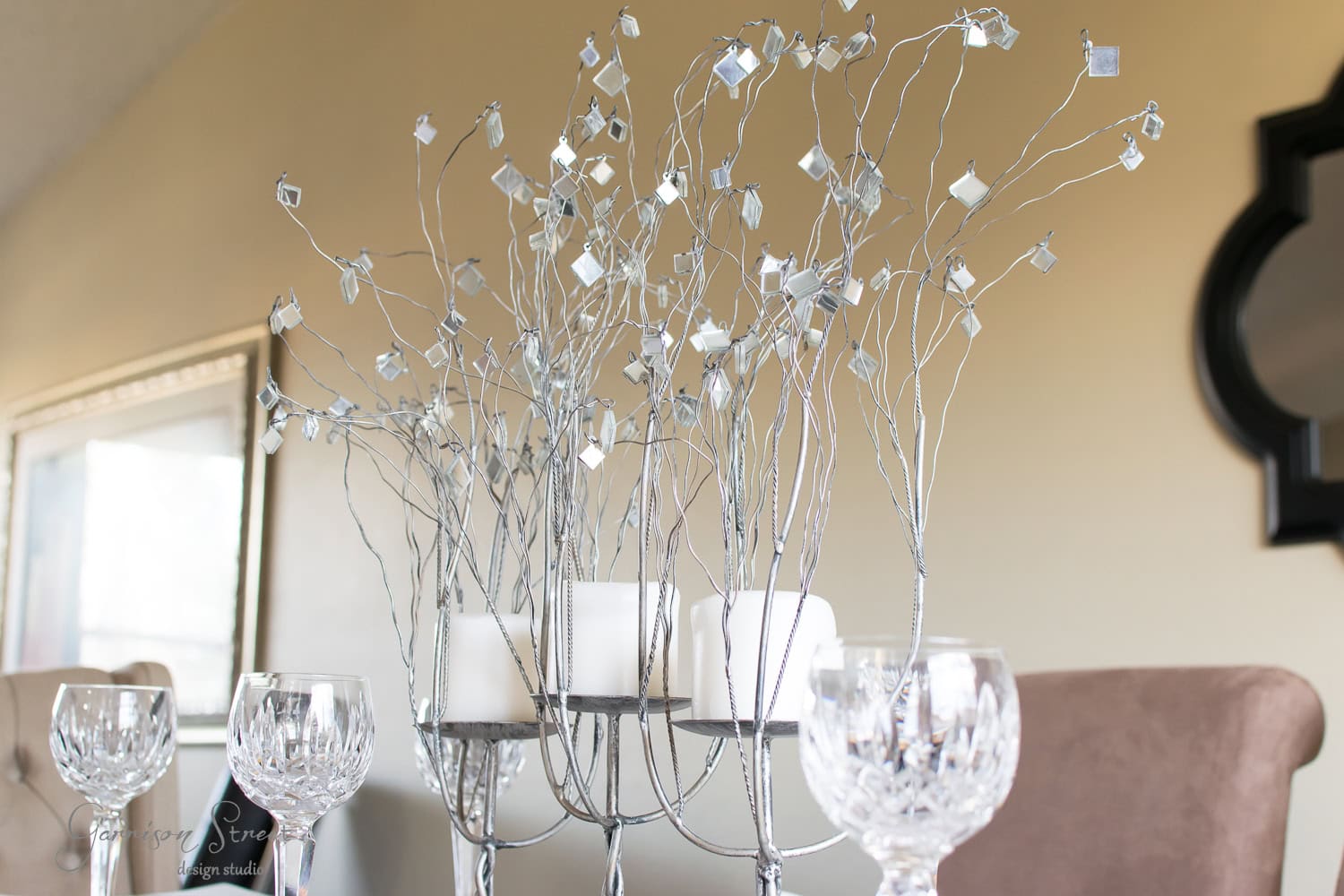 Elegant centerpiece with silver branches and small square mirrors, surrounded by white candles and crystal wine glasses on a dining table. A blurred mirror hangs on the wall in the background.