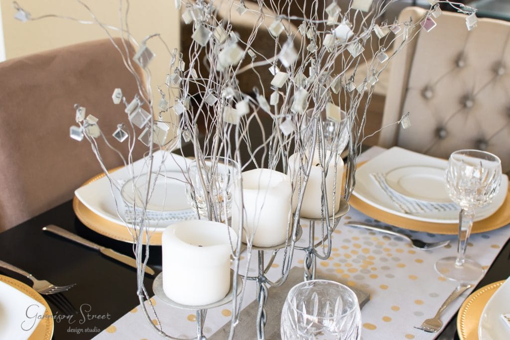 Home A dining table elegantly set with white plates, gold chargers, crystal glasses, and silverware. Centerpieces include white candles and decorative silver branches with crystal accents. The chairs have beige upholstery.