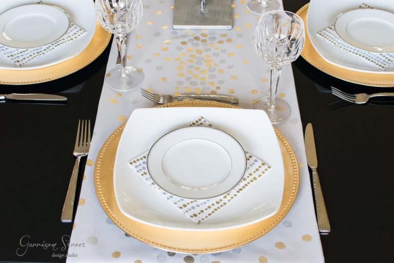 A formal dining table setting with white and gold plates on a black table. Each setting includes a crystal wine glass, silver cutlery, and a dotted napkin. The table runner has a gold and silver spotted design.