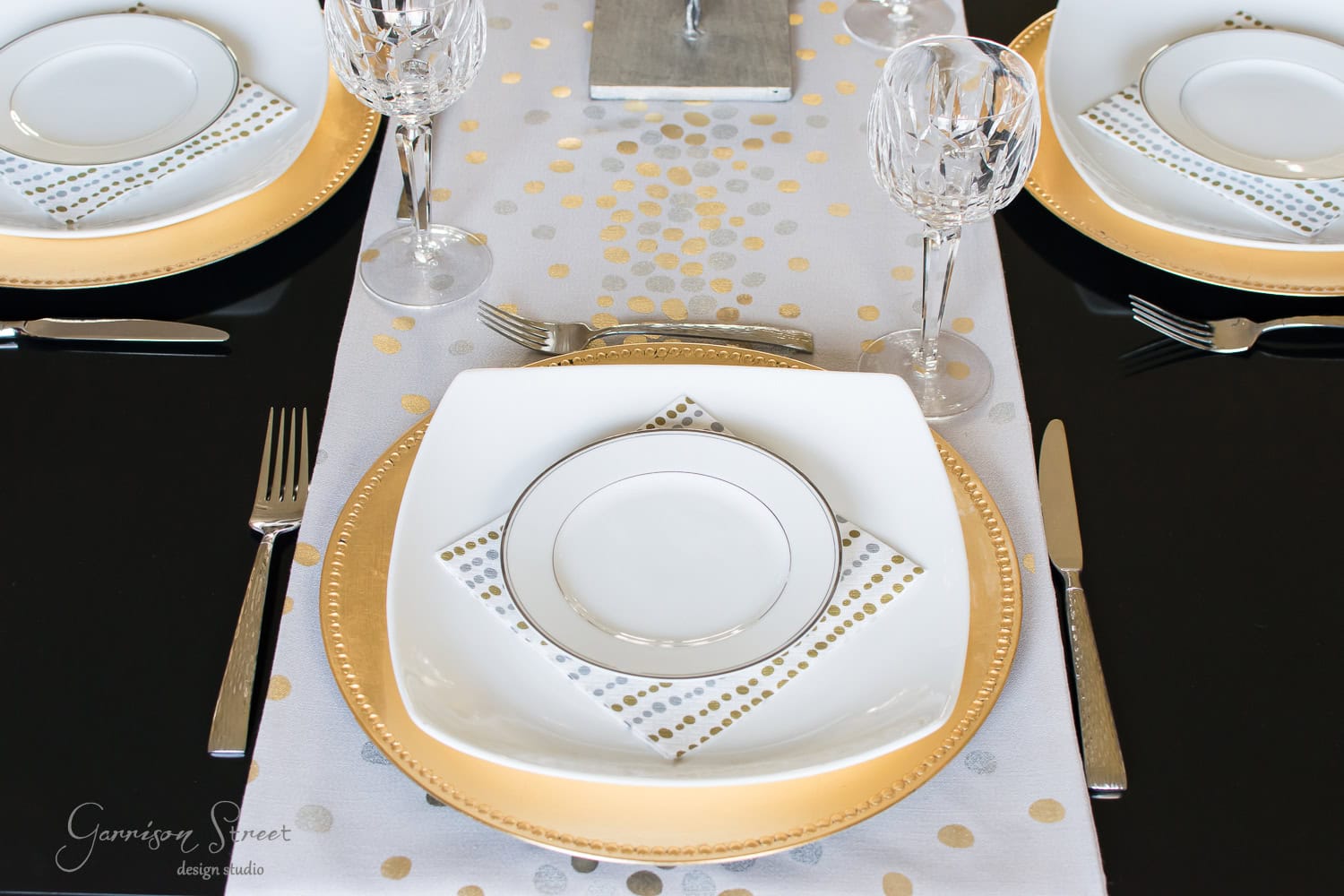 A formal dining table setting with white and gold plates on a black table. Each setting includes a crystal wine glass, silver cutlery, and a dotted napkin. The table runner has a gold and silver spotted design.