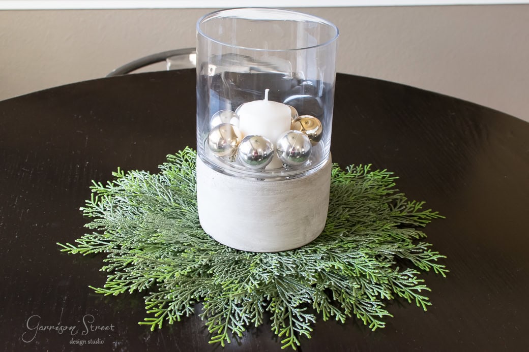 A white candle surrounded by silver ornaments is placed inside a glass and stone holder on a dark round table. Greenery is arranged around the base, creating a simple and elegant centerpiece.