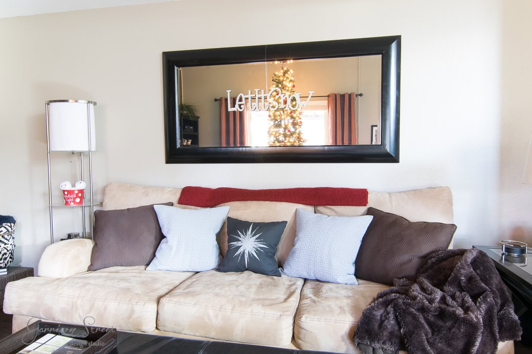 A cozy living room featuring a beige sofa with multiple cushions, a red blanket, and a dark throw. A large framed mirror reflects a lit Christmas tree. A tall lamp stands beside the sofa.