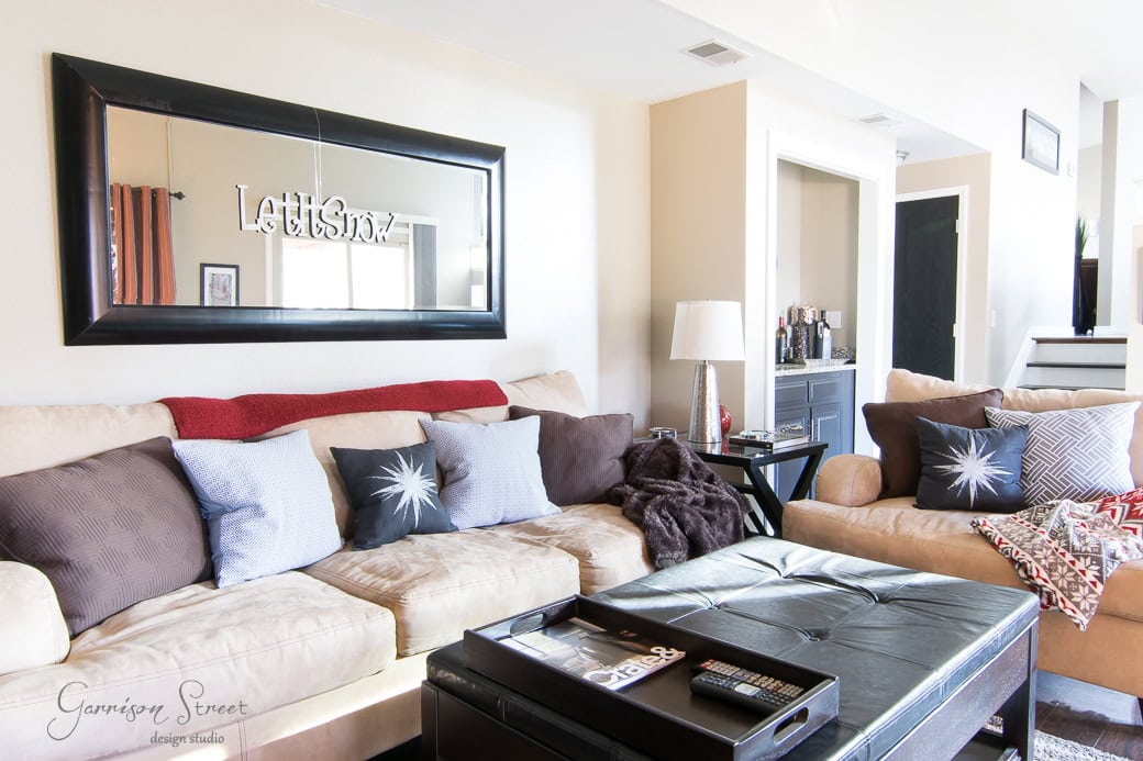 A cozy living room featuring a beige sofa adorned with assorted throw pillows and a red blanket. A large framed mirror with "Let's Stay" decor hangs above. A black ottoman serves as a coffee table, and a console table is visible in the background.