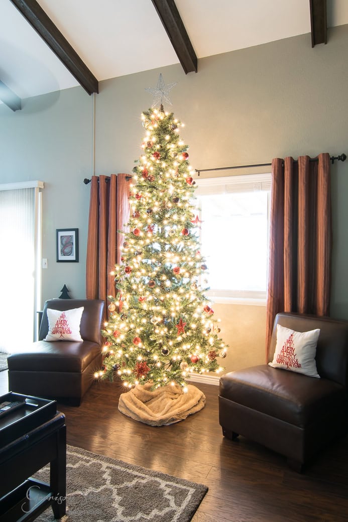 A tall, decorated Christmas tree with lights and ornaments stands by a window in a cozy living room. Two brown chairs with festive pillows are placed on either side. The room has wooden floors and beams, with curtains drawn back.