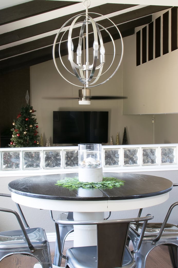 A modern dining area features a black and white round table with sleek metal chairs. A contemporary spherical chandelier hangs above. The background includes a wall-mounted TV and a small Christmas tree, with light filtering through glass blocks.
