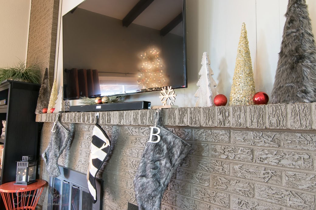 A decorated fireplace mantle with Christmas stockings hanging. Glittery trees, red ornaments, and a small snowflake decor adorn the mantle. A lit tree is reflected in the TV above. Cozy, festive holiday atmosphere.