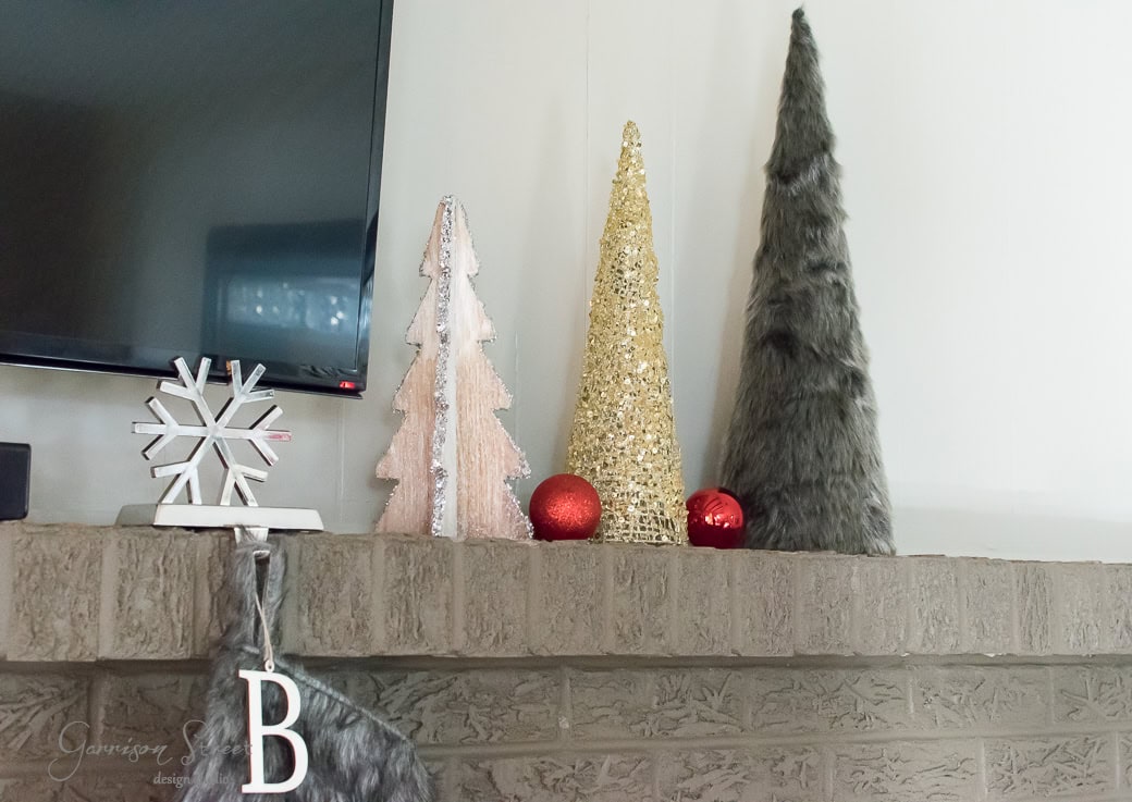 A mantel decorated with a silver snowflake, pink and gold cone-shaped trees, red ornaments, and a gray furry cone. A Christmas stocking with the letter "B" hangs over the edge. A TV is mounted on the wall beside the decorations.
