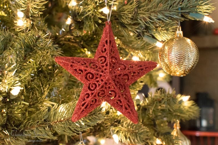 A red, ornamental star with intricate swirls hangs on a Christmas tree adorned with warm white lights and a gold bauble. The branches are lush and green, creating a festive holiday atmosphere.