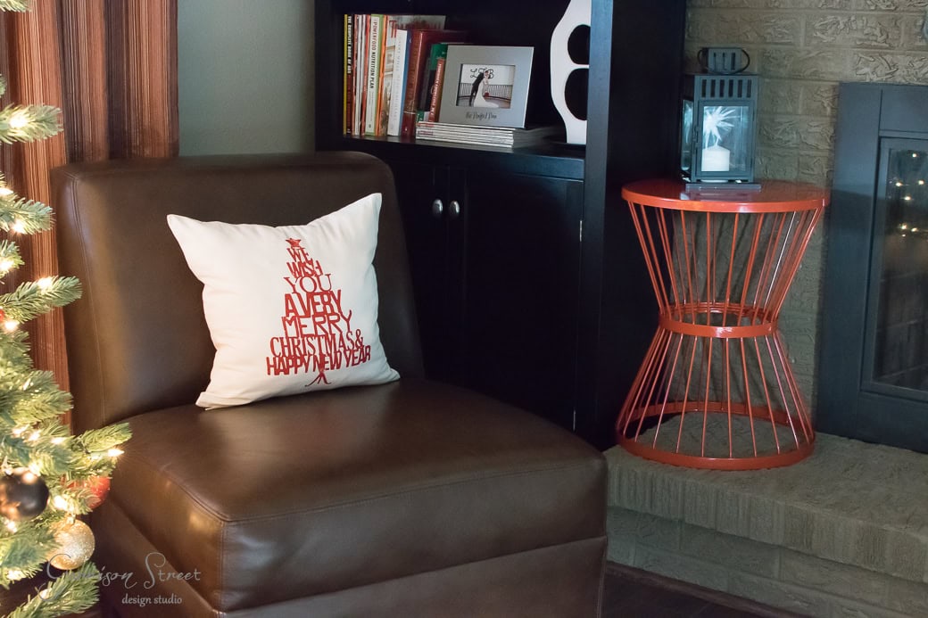 Cozy living room corner with a brown leather chair featuring a cushion that reads "We Wish You A Very Merry Christmas & Happy New Year." There's a small orange table, bookshelf, and a decorated Christmas tree nearby.
