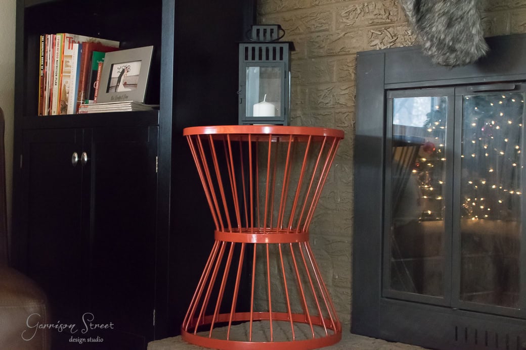 A modern living room scene featuring a red wire side table near a fireplace. On the table sits a white candle in a black lantern. Books and magazines are on a shelf in the background, and decorative lights are visible near the firewood.