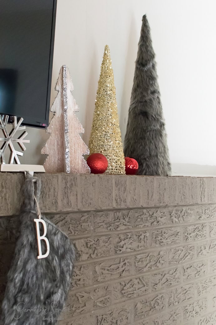 A mantel decorated with three conical trees: one silver, one gold, and one covered in gray fur. Two red ornaments sit nearby. A gray stocking with the initial "B" hangs from the mantel's edge.