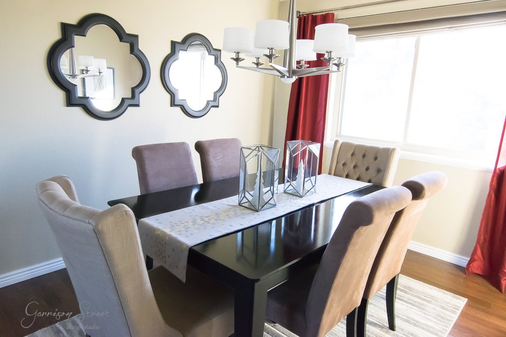 Dining room with a dark wooden table, beige upholstered chairs, and a white table runner. Two geometric candle holders on the table. Wall features two decorative mirrors. A modern chandelier hangs above, and red curtains frame a window in the background.
