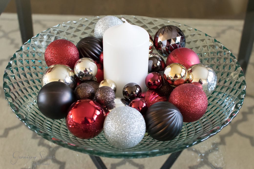 A decorative glass bowl filled with a variety of metallic and glittery Christmas ornaments in red, silver, and gold hues, surrounding a white pillar candle placed at the center.