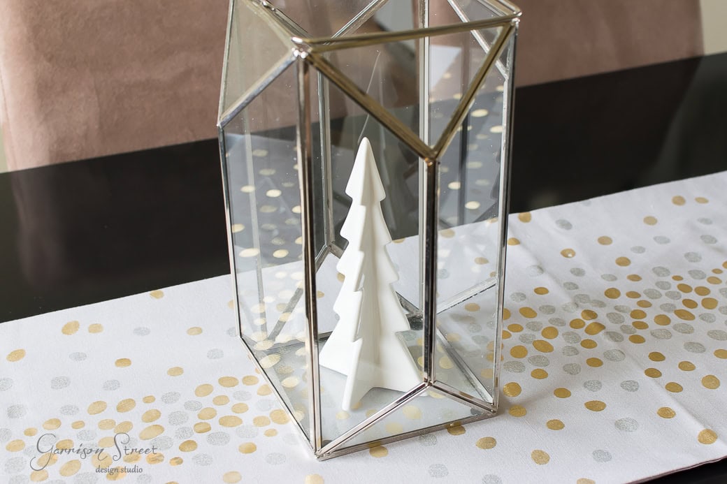 A decorative white geometric tree inside a glass polygonal terrarium sits on a table. The table runner features gold and silver dots.