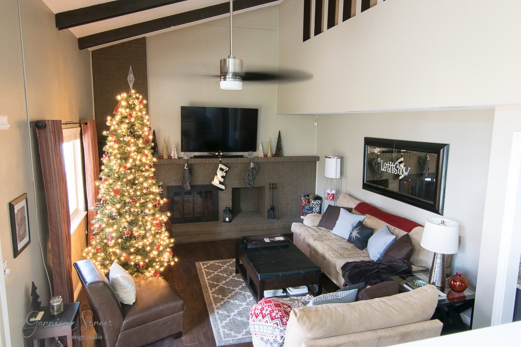 A cozy living room decorated for Christmas, featuring a tall, lit Christmas tree next to a fireplace adorned with stockings. Couches and a coffee table complete the seating area, with a large TV mounted above the mantelpiece.