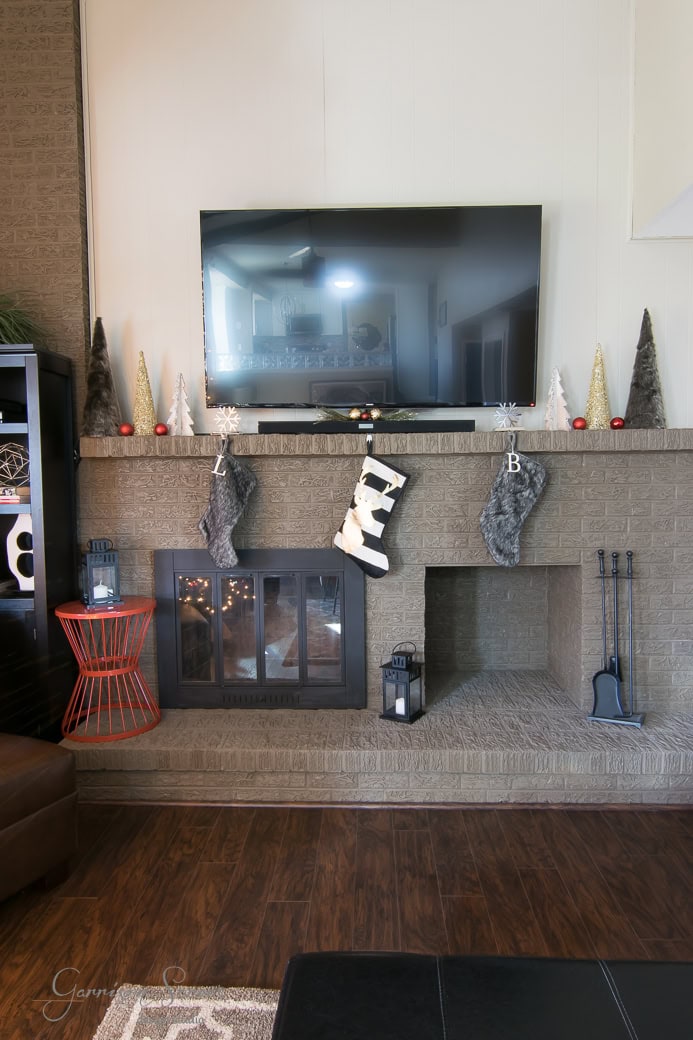 A living room with a brick fireplace, a TV mounted above, and Christmas stockings hanging from the mantel. The mantel is decorated with cone-shaped trees. A red wire side table and fireplace tools are nearby. The floor is wood.