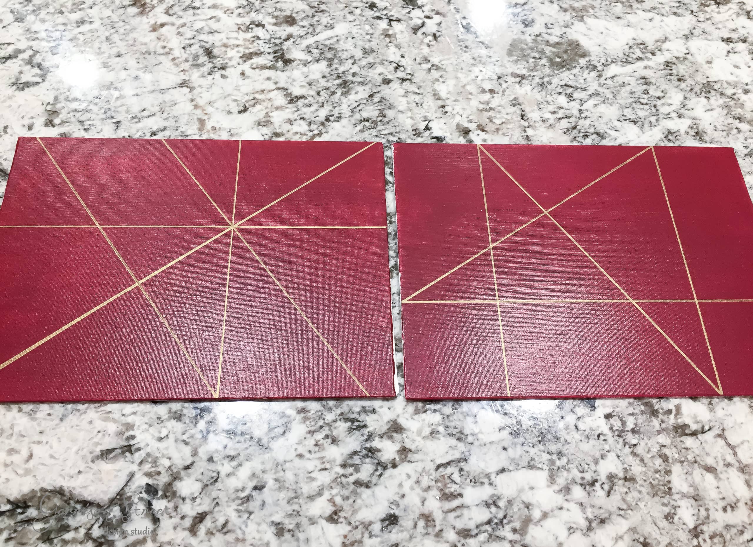 Two square red coasters with gold geometric line designs, resembling pieces of modern art, are placed side by side on a speckled granite countertop.