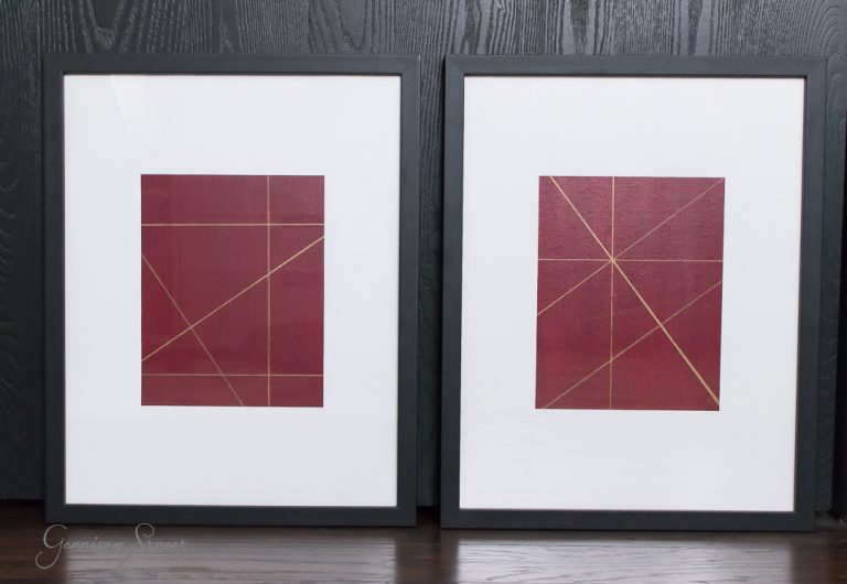 Two framed geometric abstract artworks, perfect for a DIY art enthusiast, feature intersecting gold lines on red backgrounds. The frames are black with white matting, resting gracefully against a dark wood wall.