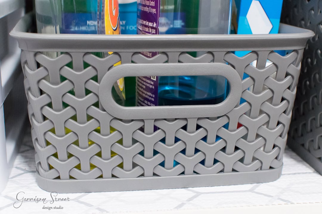 A gray plastic storage basket with a lattice pattern holds various cleaning bottles, visible through the open weave. Perfect for organization, it sits neatly on a textured paper-lined shelf beneath the kitchen sink.