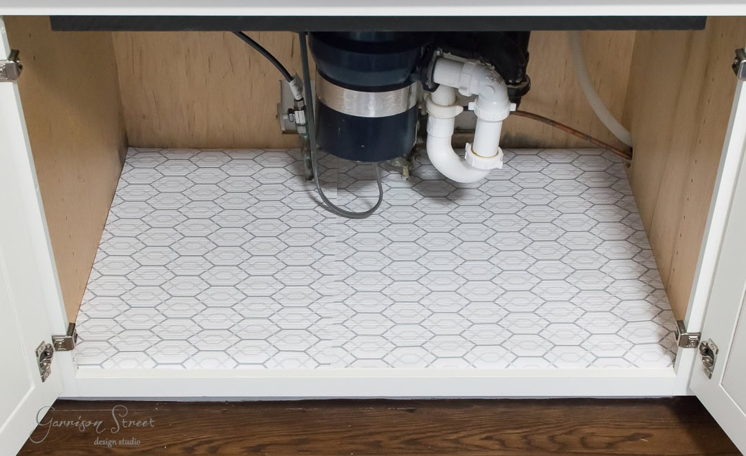 The image shows the interior of an organized kitchen cabinet beneath a sink. It features carefully arranged plumbing pipes and a garbage disposal unit. The base is lined with stylish white hexagonal-patterned tiles.