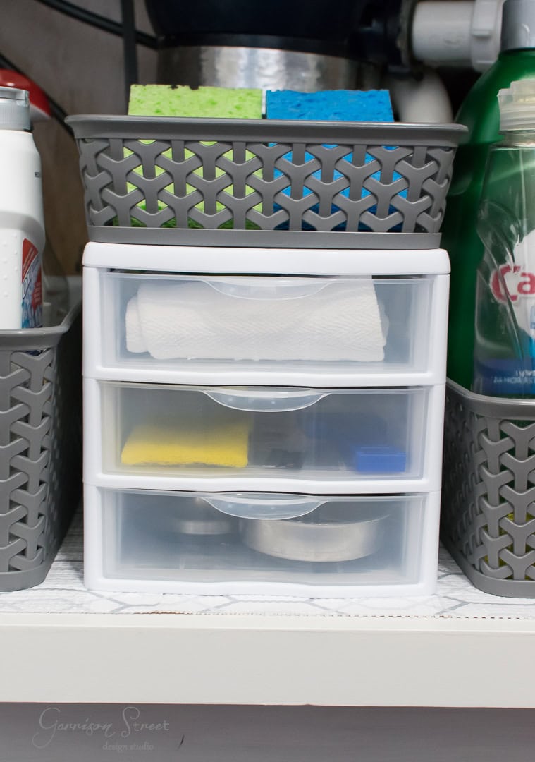A neat kitchen storage setup under the kitchen sink features a white plastic drawer organizer containing paper towels and cleaning supplies. Gray baskets on either side hold bottles and colorful sponges, ensuring excellent organization.