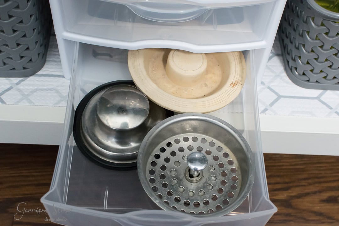An open plastic drawer showcasing a few metal and plastic kitchen sink strainers highlights the essence of organization. The background features a patterned shelf liner, with textured containers nearby, adding a touch of order to any culinary space.