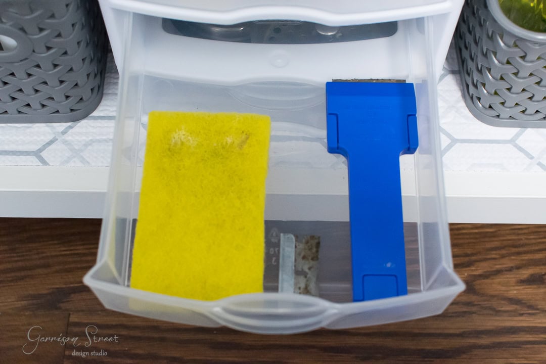 An open drawer, perfect for kitchen sink organization, holds a yellow sponge and a blue scraper. The partially transparent white drawer is complemented by woven grey baskets nearby, all resting on a wooden surface.