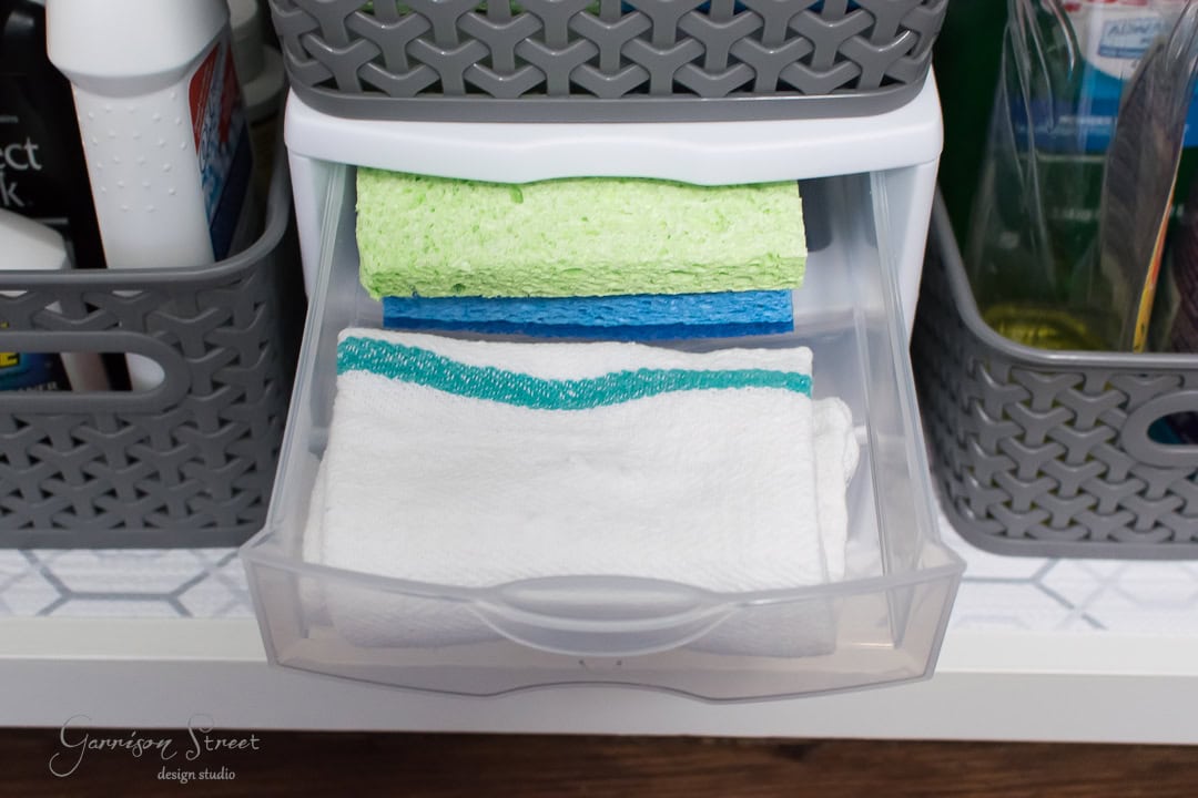 A plastic drawer opens to reveal two neatly folded cleaning cloths, one green and one white with blue trim, perfectly organized for easy access by the kitchen sink. They are part of a meticulously arranged cleaning supply setup, surrounded by gray baskets.