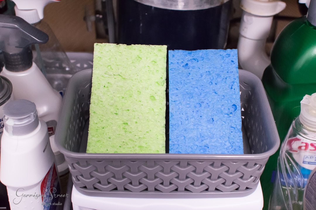 Two sponges, one green and one blue, are placed vertically in a gray basket near the kitchen sink. The basket is surrounded by various cleaning supplies, including bottles and containers, promoting tidy organization on the shelf.