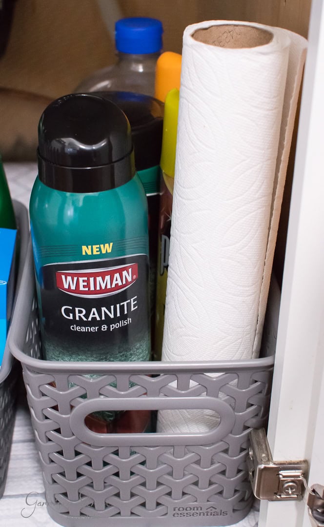 A storage basket for organization, containing a granite cleaner spray and a roll of paper towels, is neatly placed inside the cabinet under the kitchen sink. The basket is gray with perforated sides, and nearby, a blue bottle is partially visible.