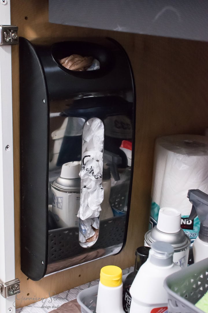 A wall-mounted grocery bag dispenser brimming with plastic bags hangs neatly above shelves that reveal a well-organized array of household supplies. Spray cans, paper towels, and bottles are methodically sorted in plastic bins, showcasing meticulous organization in this utility area.