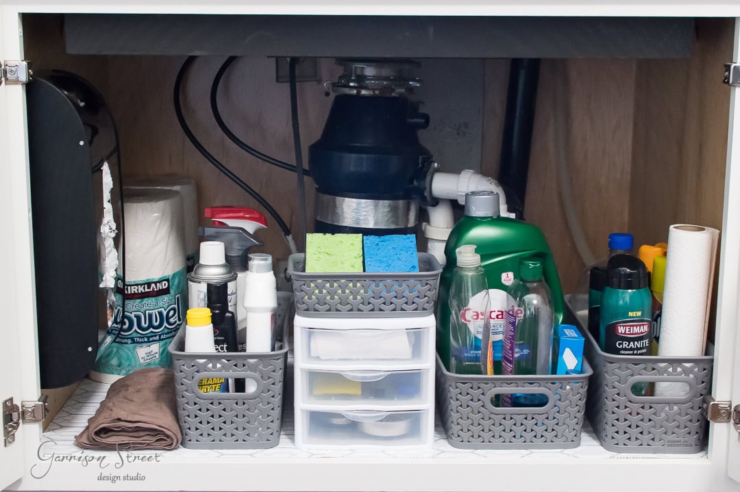 Under-sink cabinet neatly organized with cleaning supplies. Baskets contain sponges, bottles, and containers of detergent. Paper towels are on one side, and a garbage disposal unit is visible in the back.