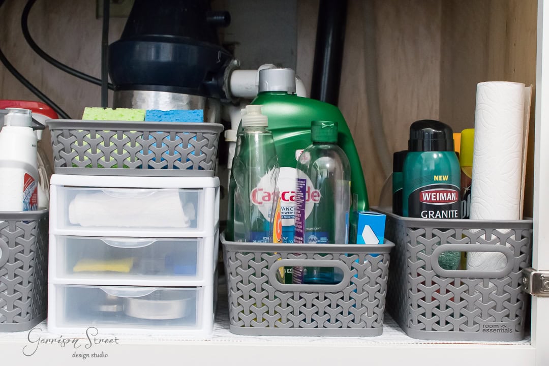 Under the kitchen sink, you'll find an impeccably organized cabinet with cleaning supplies neatly arranged. Sponges, cleaning products, paper towels, and plastic storage baskets are all in their perfect place.
