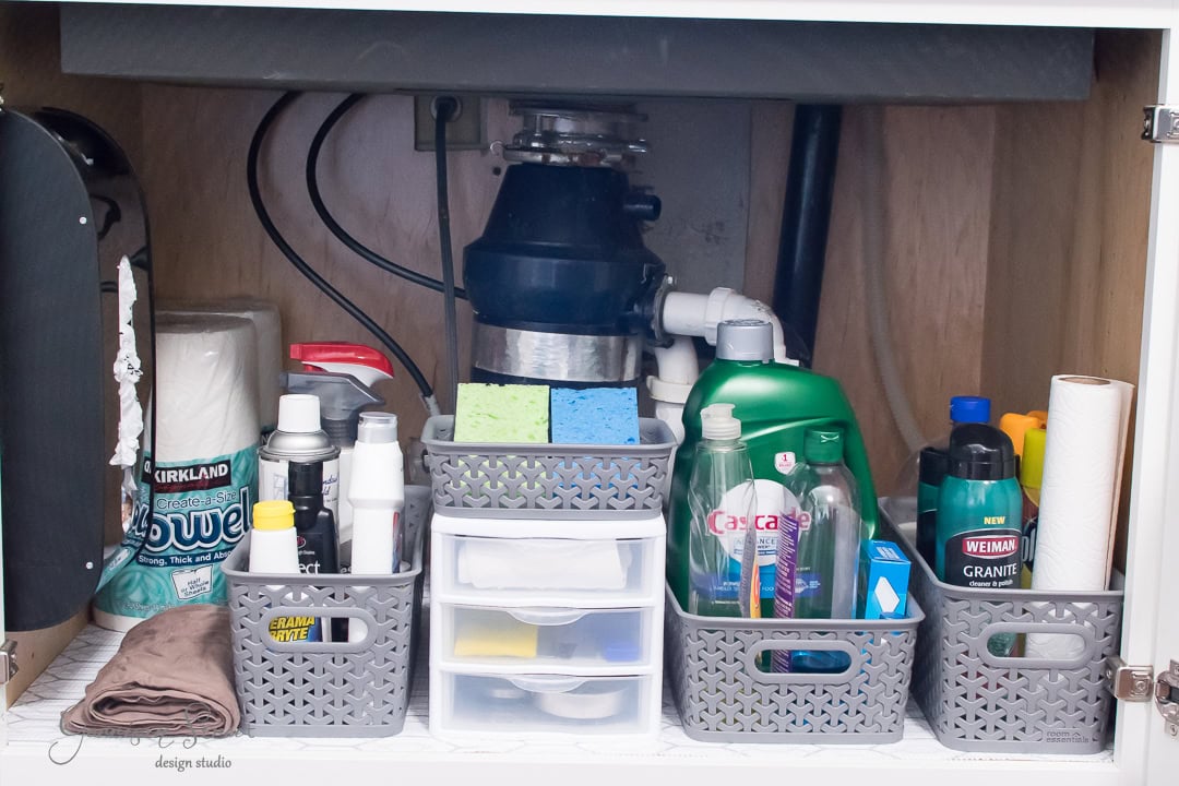 Organized under-sink cabinet with assorted cleaning supplies exemplifies kitchen sink organization. Three baskets hold bottles, sponges, and rolls, while a central drawer unit contains small items. A garbage disposal unit is visible in the background.