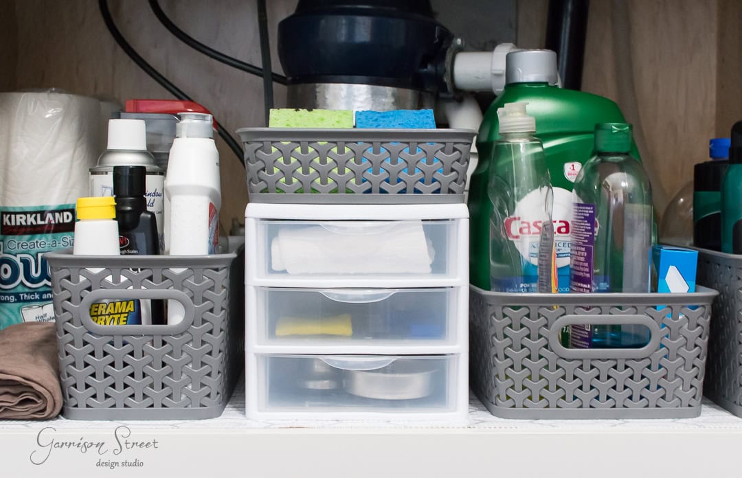 A neatly organized kitchen sink storage area features grey baskets holding cleaning supplies, sponges, and bottles of detergent. A small plastic drawer unit in the center keeps various items tidy. Pipes and a garbage disposal unit are visible at the back, showcasing efficient organization.