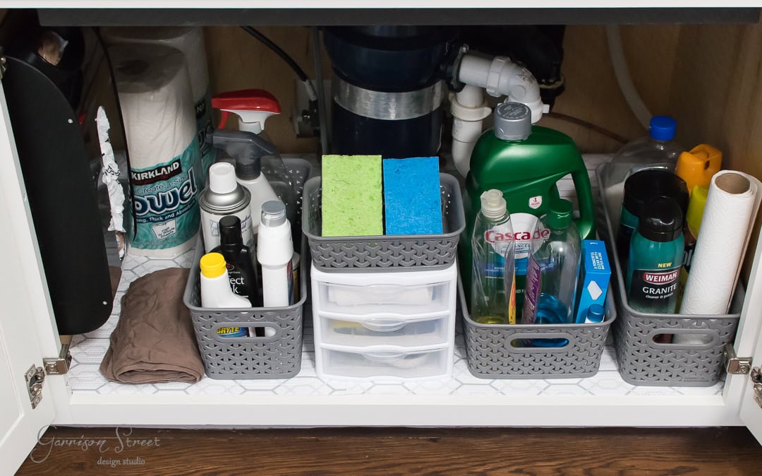 Under the kitchen sink, an organized cabinet features baskets holding cleaning supplies, sponges, and paper towels. Dish soap, spray bottles, and a garbage disposal unit are visible. A drawer neatly organizes smaller items, while decorative paper lines the cabinet for a stylish touch..